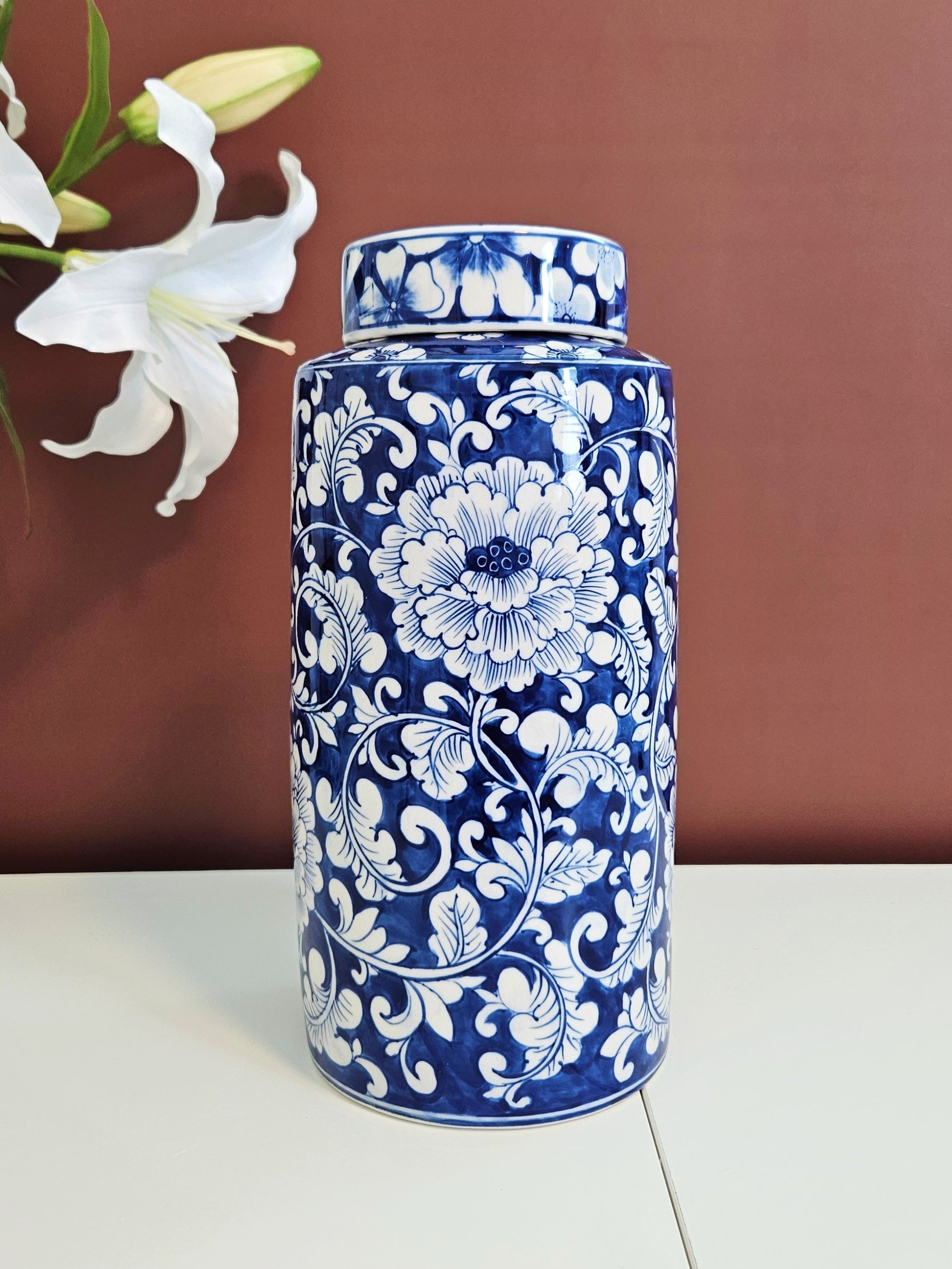 A tall cylindrical blue and white chinoiserie porcelain lidded jar with an intricate floral and scroll pattern, set on a white surface against a warm brown background with a white lily flower in the upper left corner.