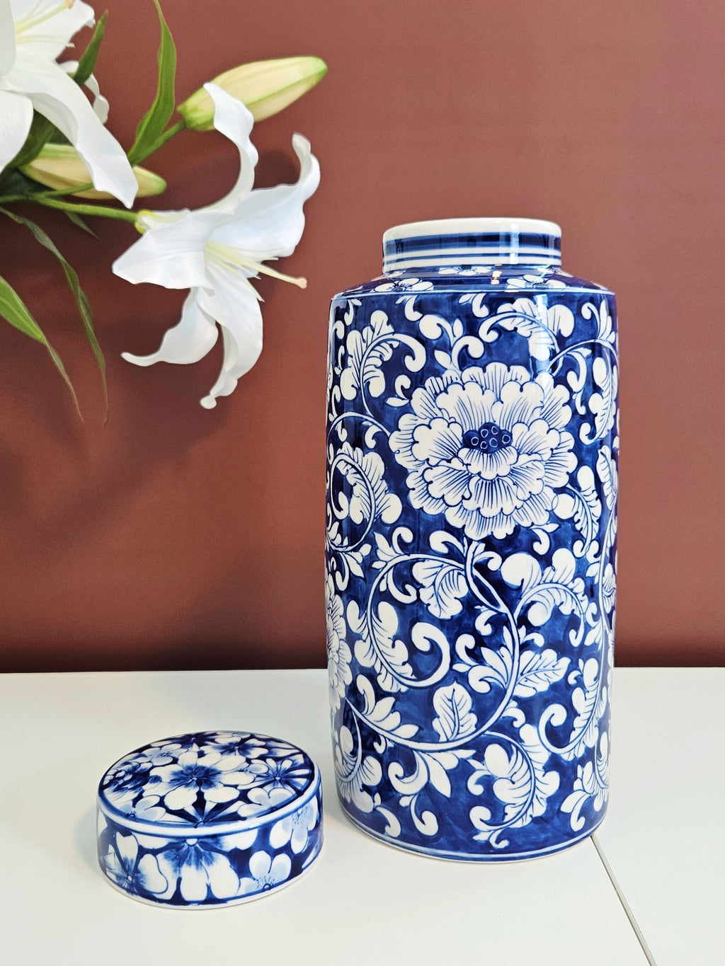 A blue and white chinoiserie porcelain storage jar with a matching lid, decorated with a floral chrysanthemum pattern, set against a terracotta background with white lilies in the corner.