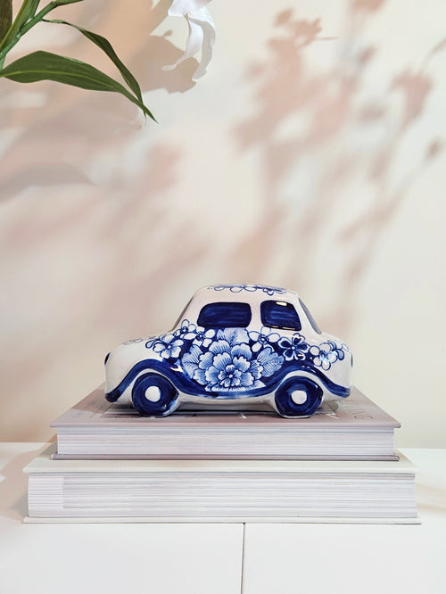 A decorative ceramic car figurine with blue and white floral patterns on a white base, displayed on top of two stacked books against a softly lit cream wall with leaf shadows.