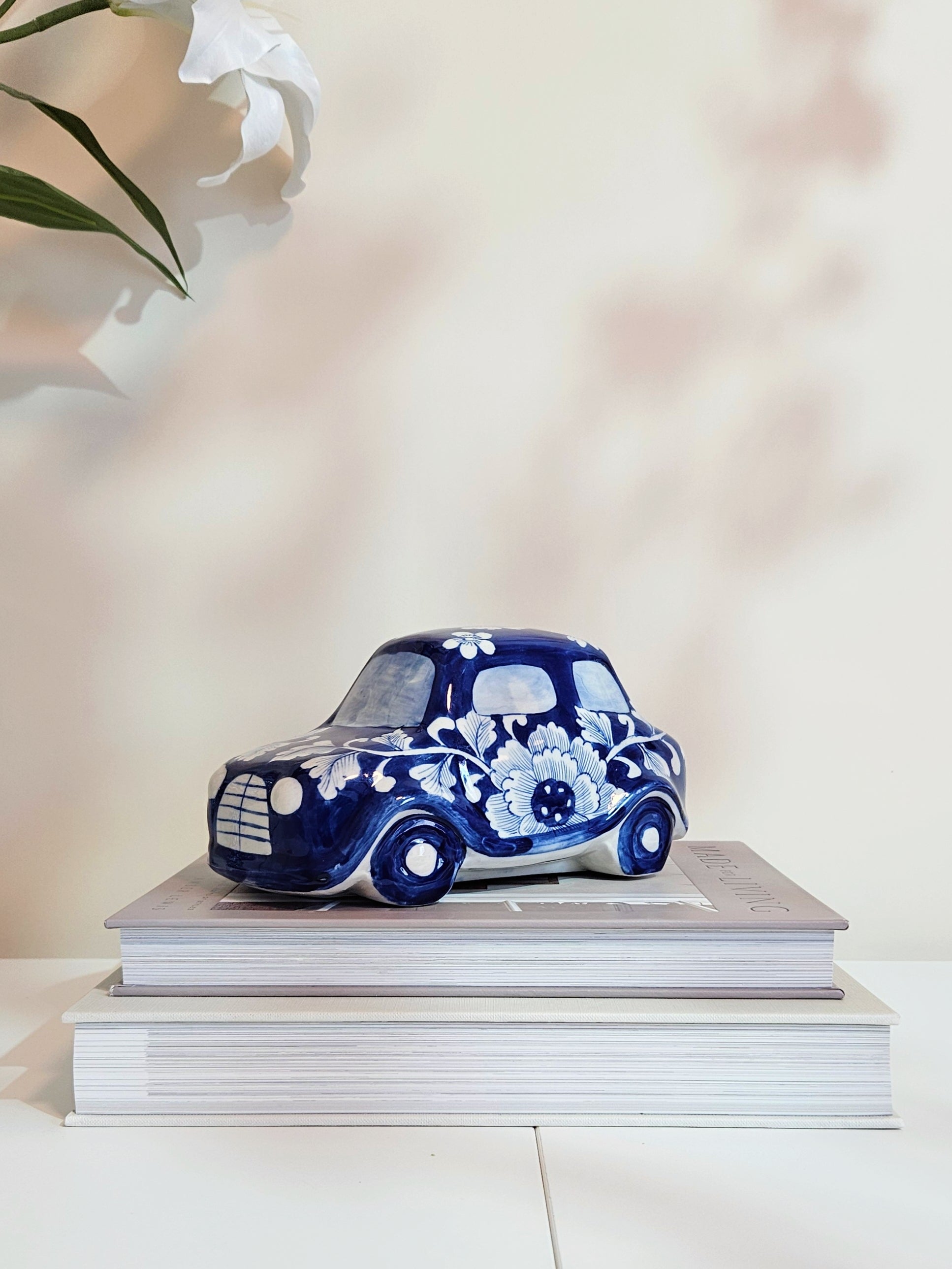 A chinoiserie decorative porcelain car figurine in deep cobalt blue with white floral patterns, displayed on top of two stacked books against a soft beige background with a blurred white flower in the upper left.