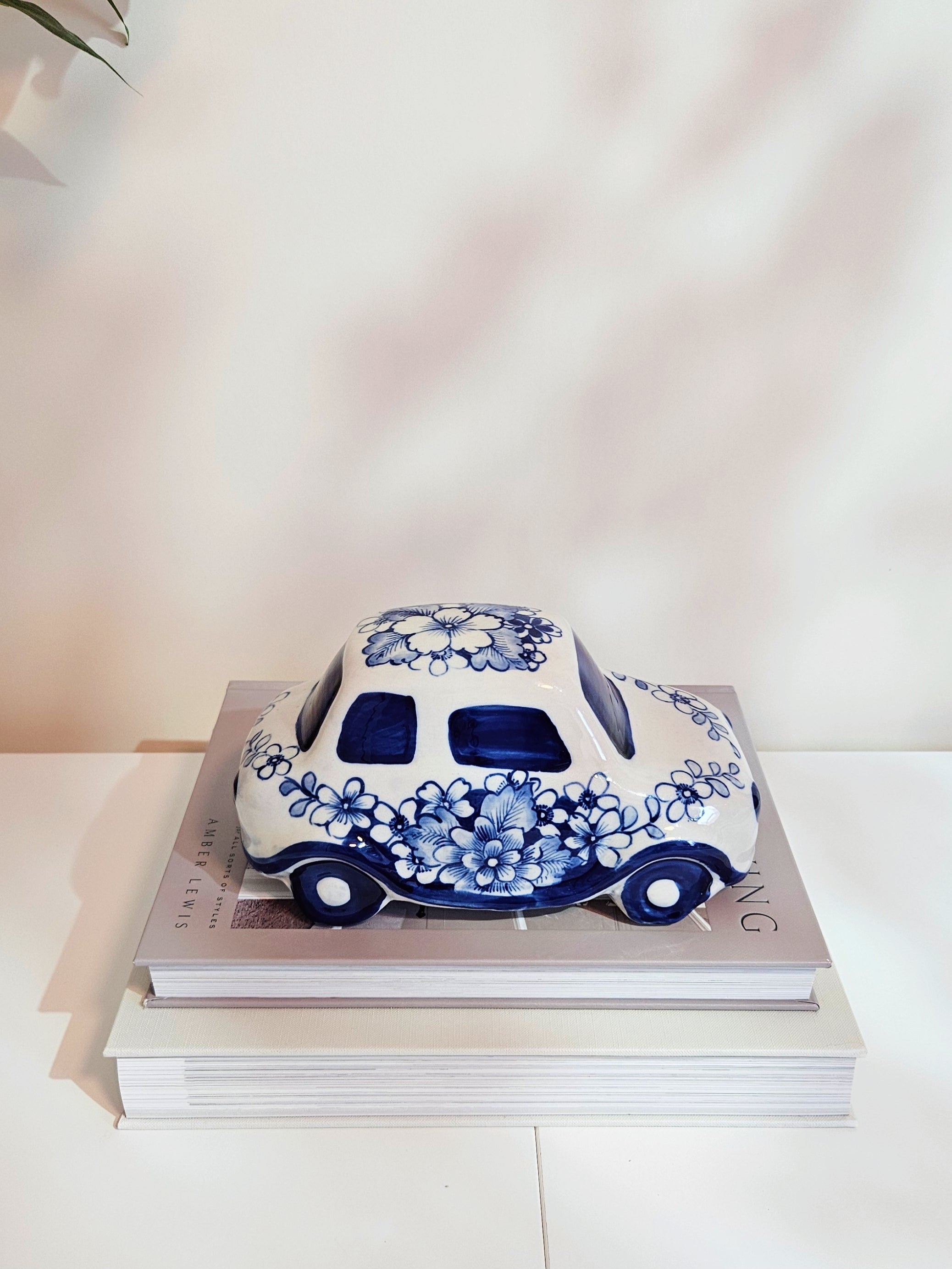 A decorative ceramic car figurine with a white and blue floral patterns, displayed on top of two stacked books against a light beige wall, showing details on the top of the car.