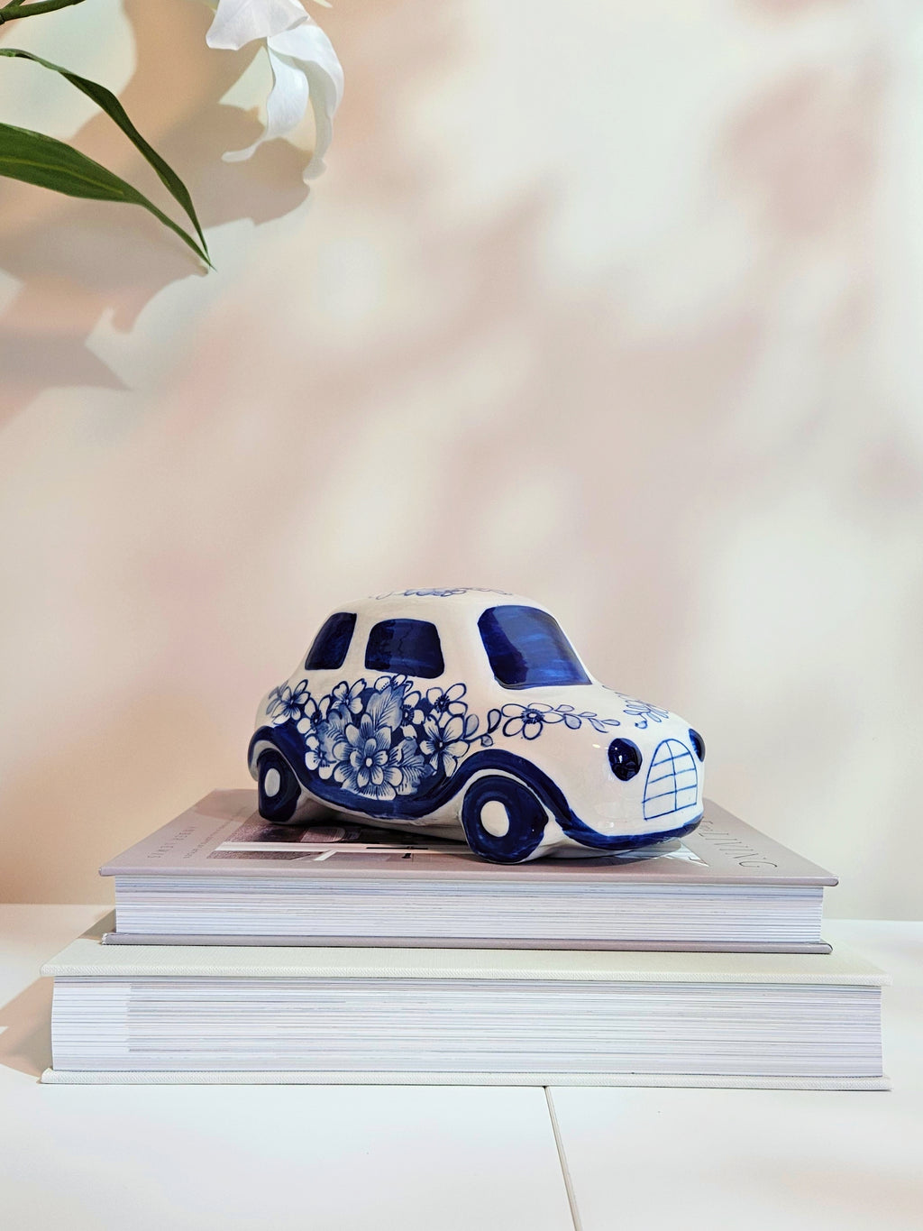 A small decorative ceramic car figurine in white and blue, styled on top of two stacked books against a soft, warm beige background with a blurred white flower in the upper left corner.