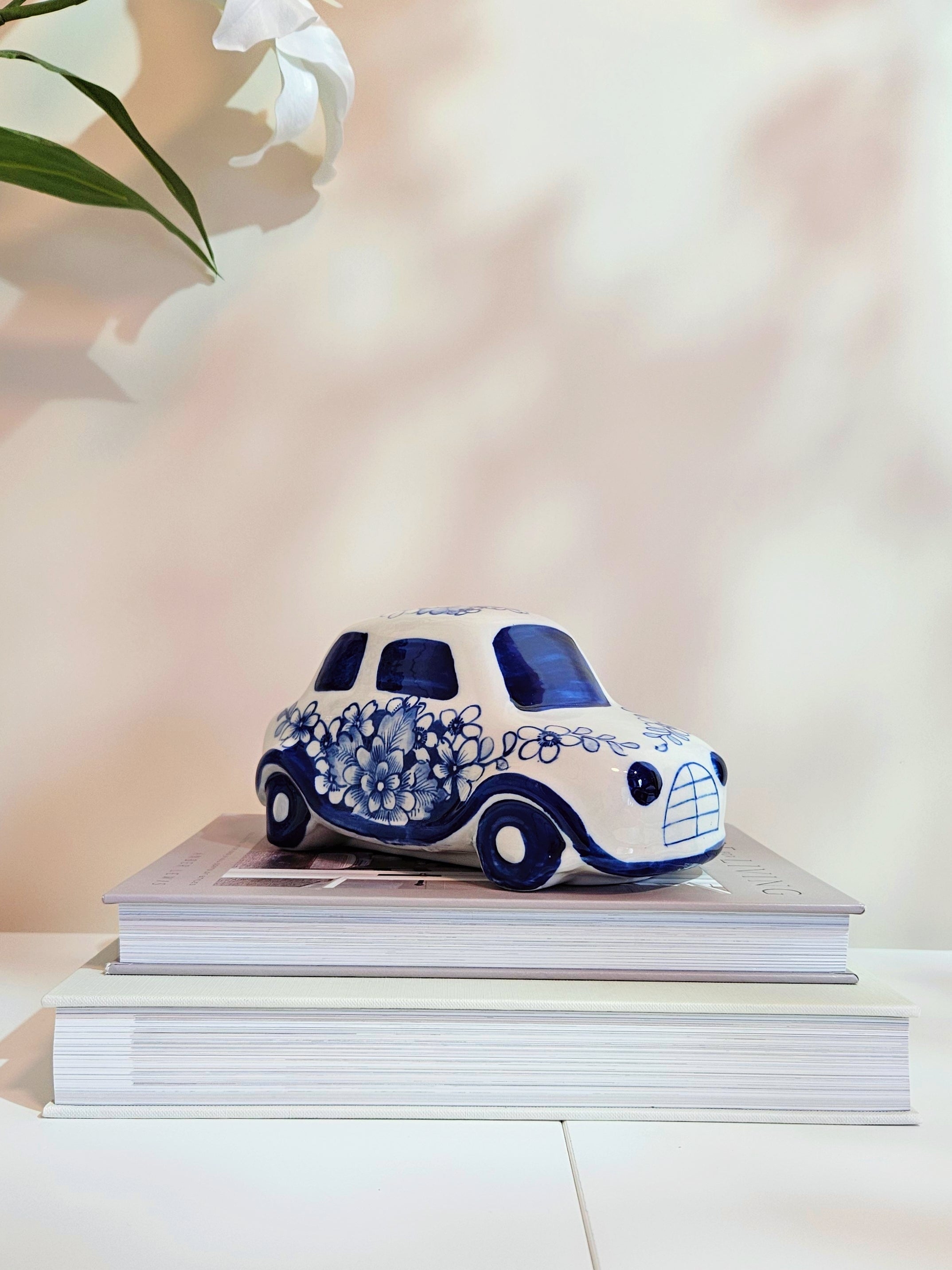 A small decorative ceramic car figurine in white and blue, styled on top of two stacked books against a soft, warm beige background with a blurred white flower in the upper left corner.