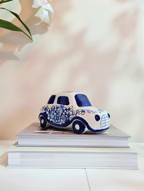 A small decorative ceramic car figurine in white and blue, styled on top of two stacked books against a soft, warm beige background with a blurred white flower in the upper left corner.