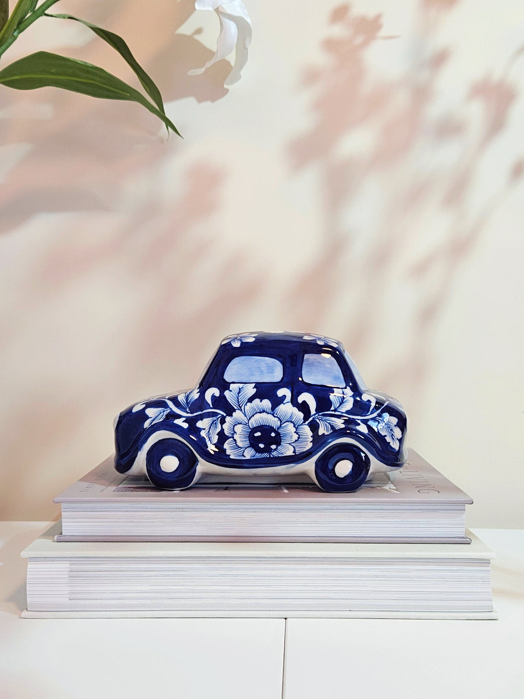 A decorative ceramic car figurine in a deep blue and white floral pattern, displayed on top of two stacked white books against a soft beige wall with leaf shadows.
