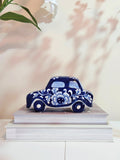 A decorative ceramic car figurine in a deep blue and white floral pattern, displayed on top of two stacked white books against a soft beige wall with leaf shadows.