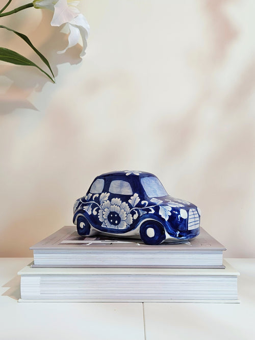 A small decorative porcelain car in a cobalt blue and white floral pattern, displayed on top of two stacked white books against a soft beige background.
