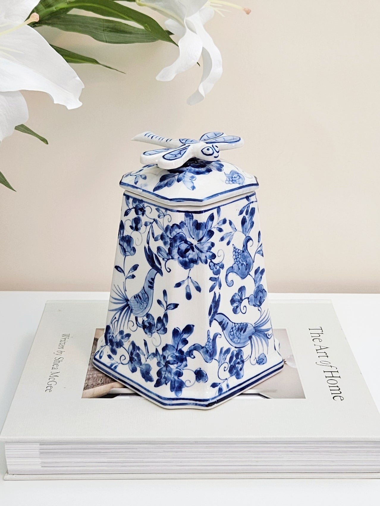 A blue and white porcelain jar with dragonfly lid featuring floral and bird patterns, displayed on top of a white book with white lilies in the background.