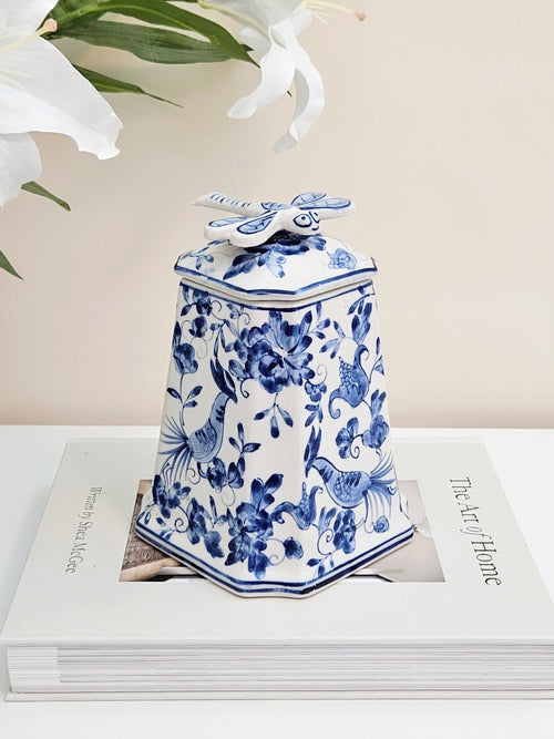 A blue and white porcelain jar with dragonfly lid featuring floral and bird patterns, displayed on top of a white book with white lilies in the background.