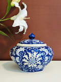 A blue and white chinoiserie porcelain lidded jar with intricate floral scrollwork, displayed on a white surface against a warm brown background with a white lily flower in the upper left.