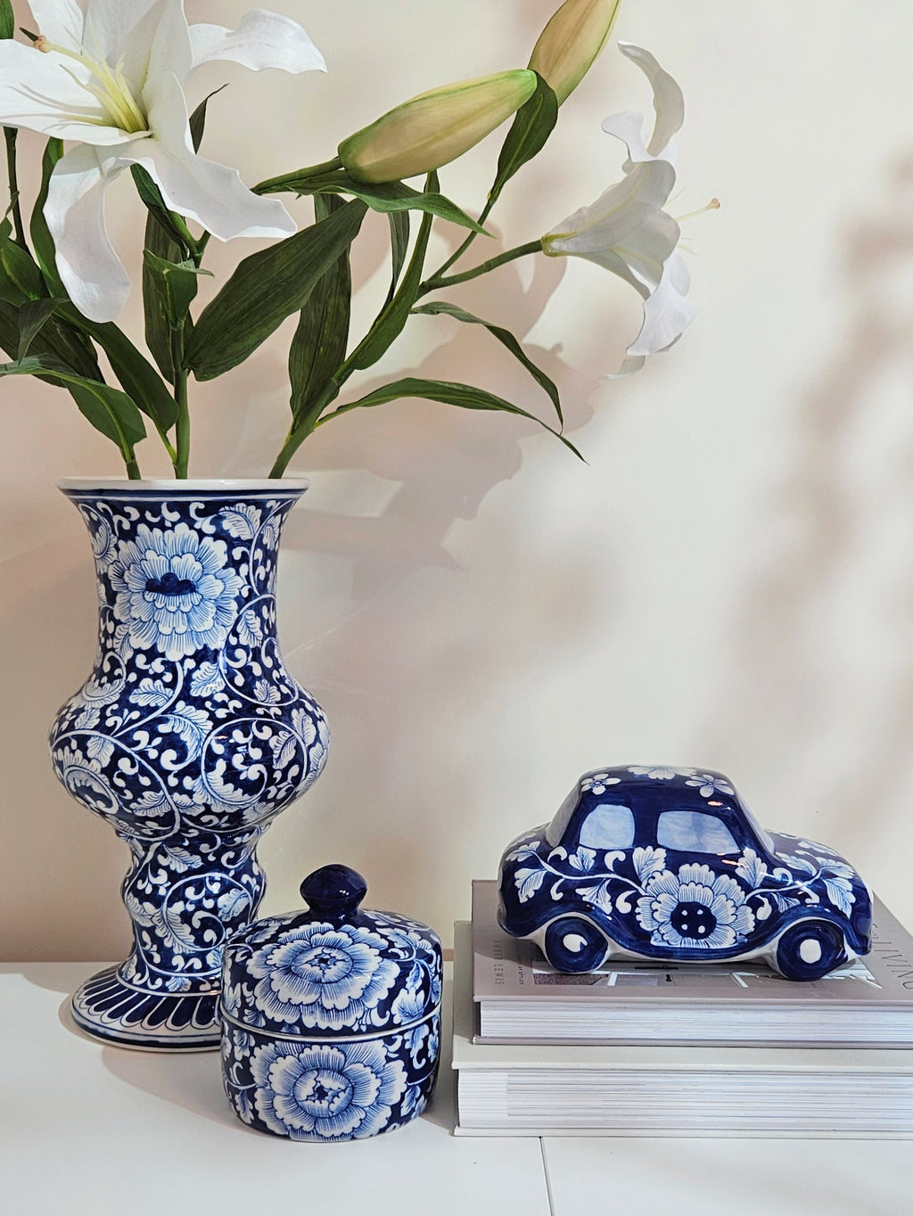 Blue and white floral porcelain ceramic set including a tall vase with white lilies, a small lidded box, and a decorative blue and white car figurine on stacked books.