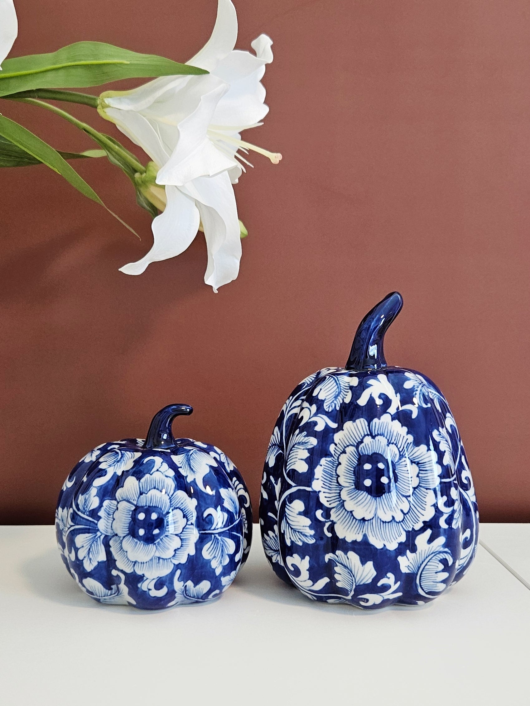 Two decorative porcelain pumpkins with a blue and white floral pattern, chinoiserie style, played on a white surface against a terracotta background with a white lily flower in the upper left.