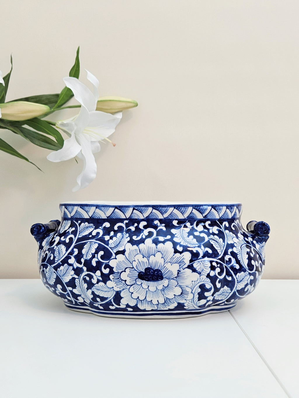 A blue and white chinoiserie porcelain decorative planter pot with floral patterns and side handles, displayed on a white surface against a light beige background with a white lily flower in the upper left corner.