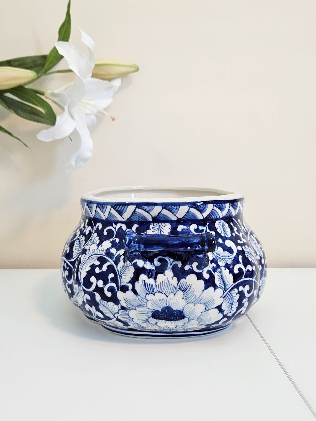 A round blue and white porcelain planter pot with a floral pattern, displayed on a white surface against a light beige background with a white lily flower in the upper left corner.