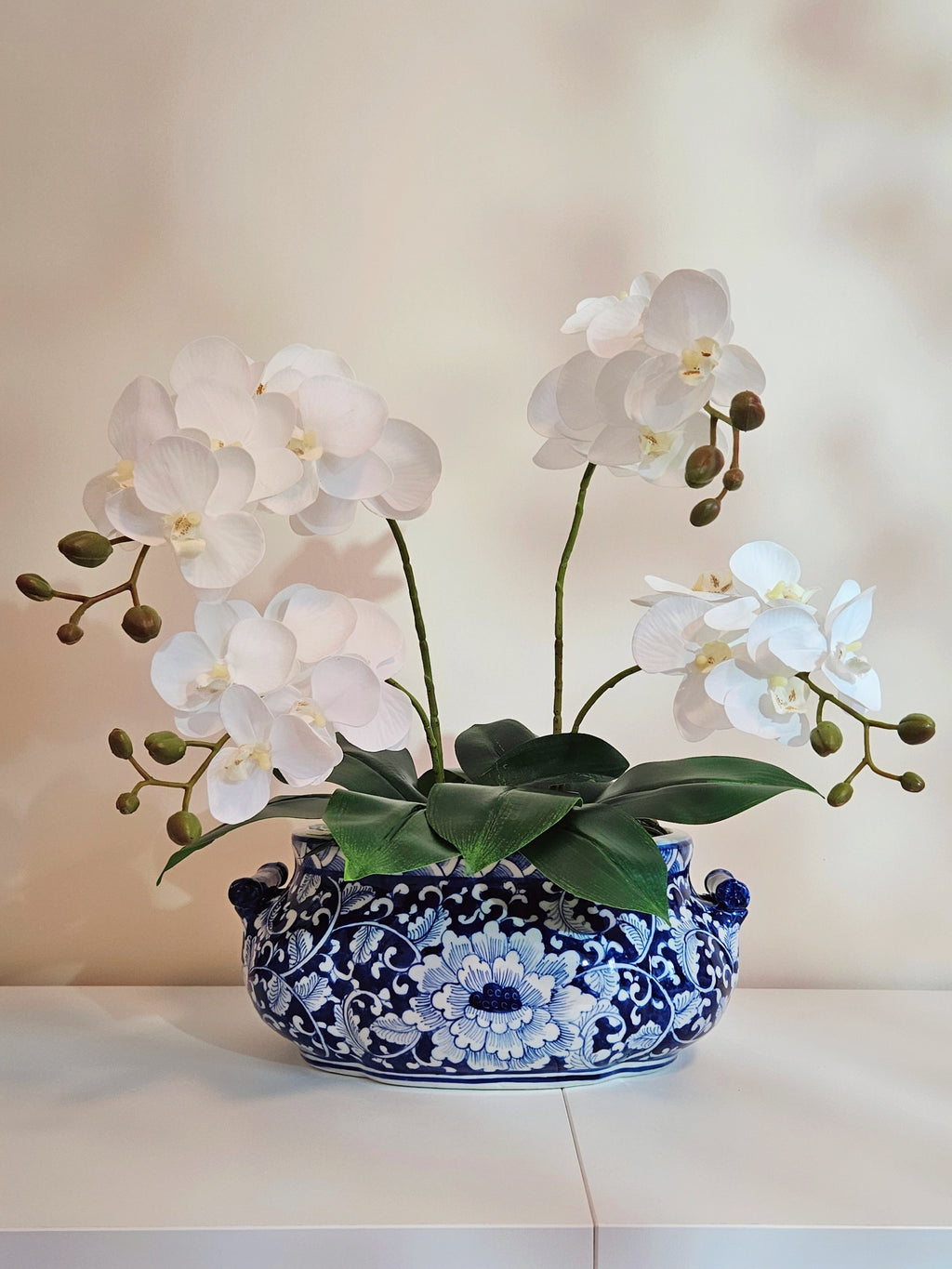 a blue and white chinoiserie ceramic planter pot with orchid displaying on a white table with beige background