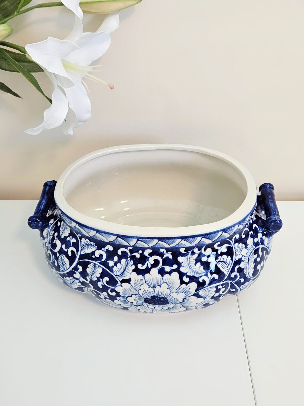 A blue and white porcelain planter pot with ornate floral patterns and side handles, displayed on a white surface with white lilies in the background.