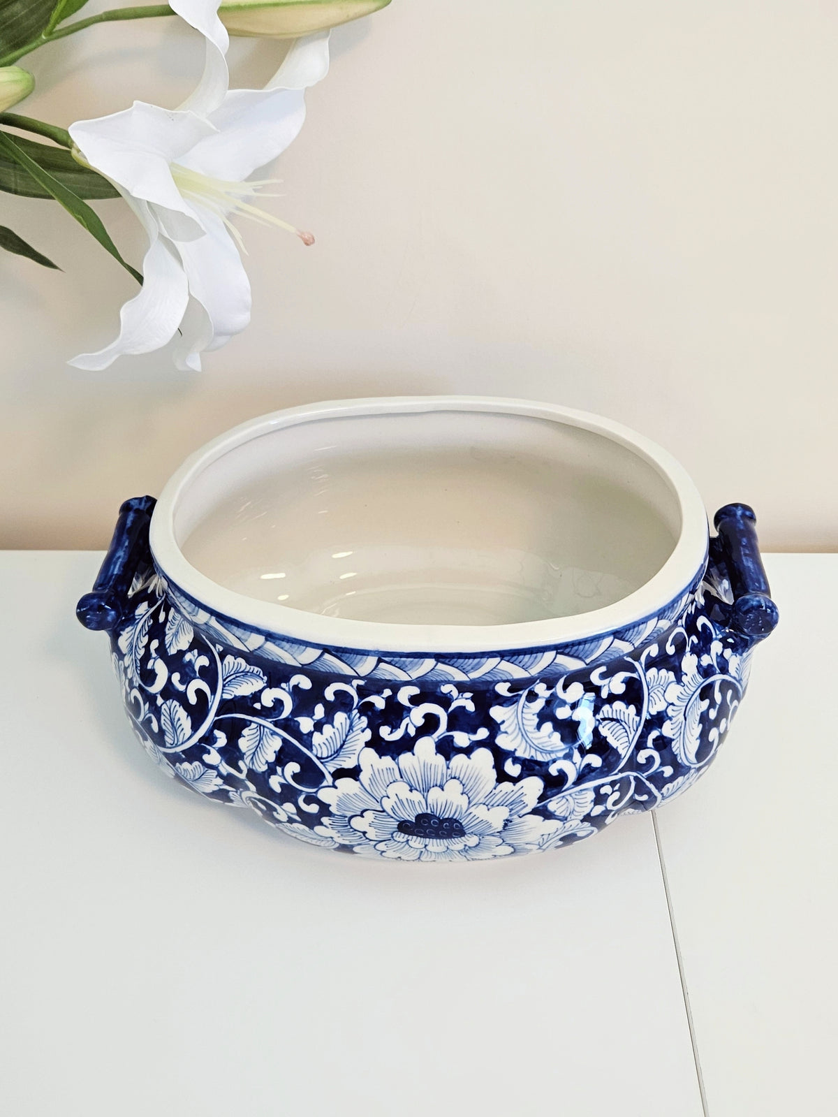 A blue and white porcelain planter pot with ornate floral patterns and side handles, displayed on a white surface with white lilies in the background.