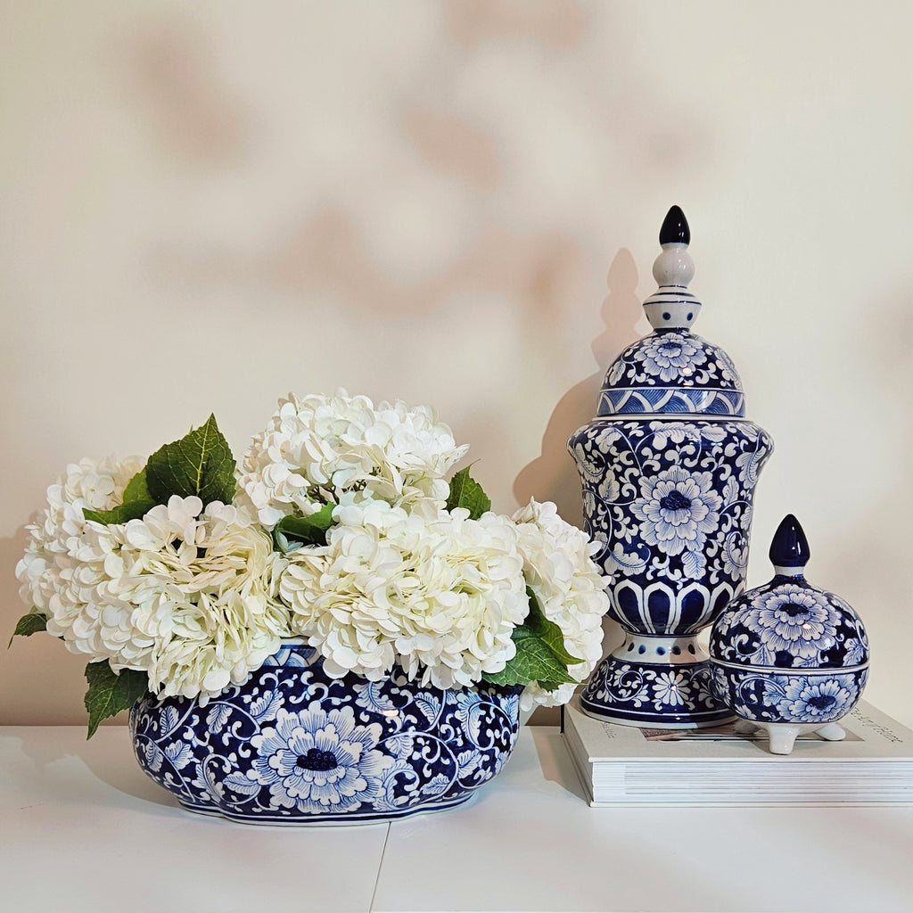 Blue and white floral ceramic decorative pieces, including a round vase filled with white flowers and two lidded jars, arranged on a white surface against a soft beige background.