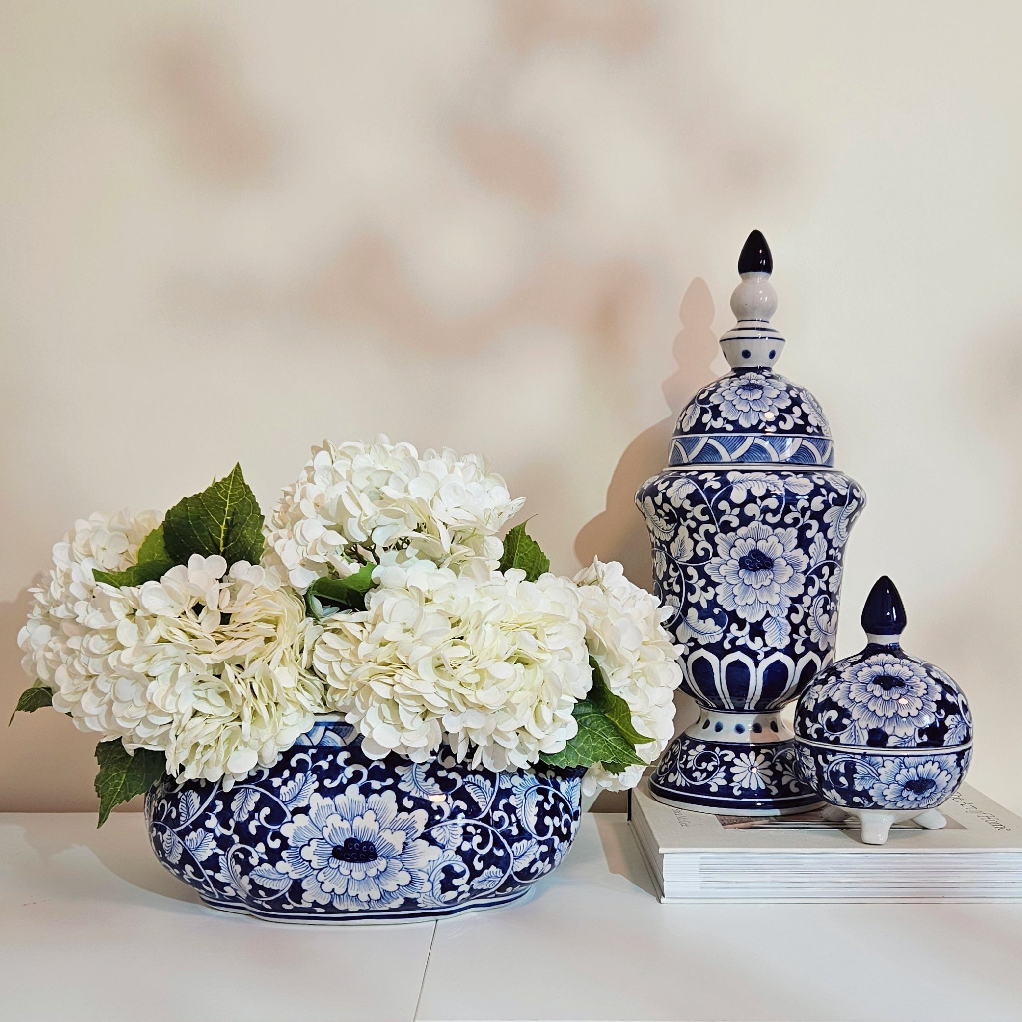 Blue and white floral ceramic decorative pieces, including a round vase filled with white flowers and two lidded jars, arranged on a white surface against a soft beige background.