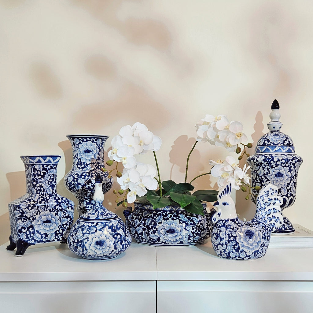 A collection of blue and white porcelain decorative vases and a lidded urn displayed on a white cabinet against a beige wall, with white orchids in the center vase.