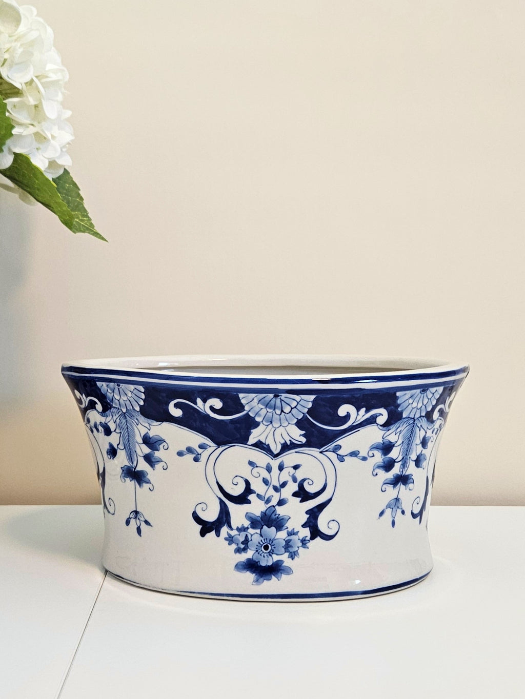 A white ceramic planter pot with a blue floral and scroll pattern, sitting on a white surface against a light beige background with white flowers in the top-left corner.