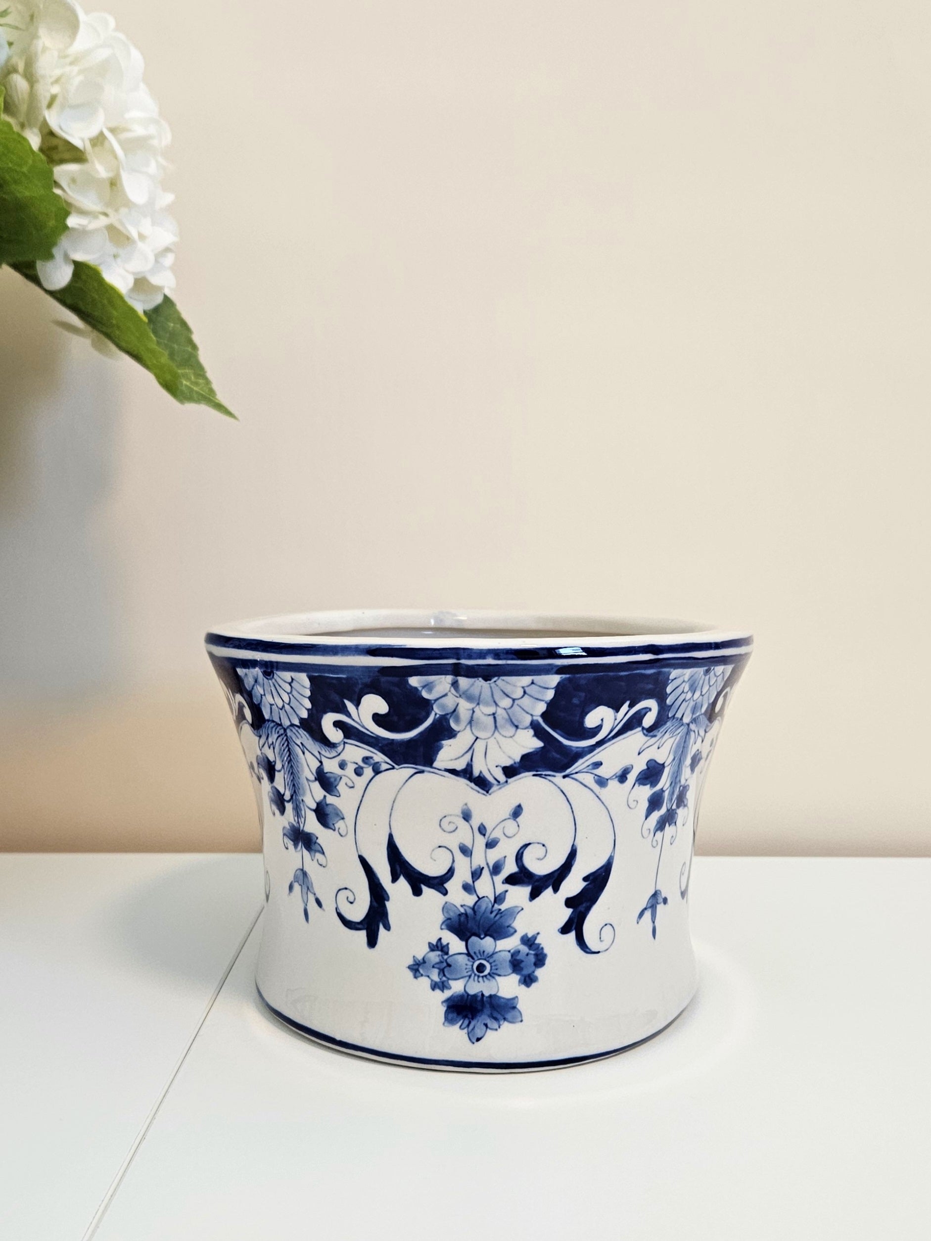 A blue and white ceramic planter pot decorated with a classic blue floral and scroll pattern, sitting on a white surface against a beige wall with a white flower in the upper left corner.