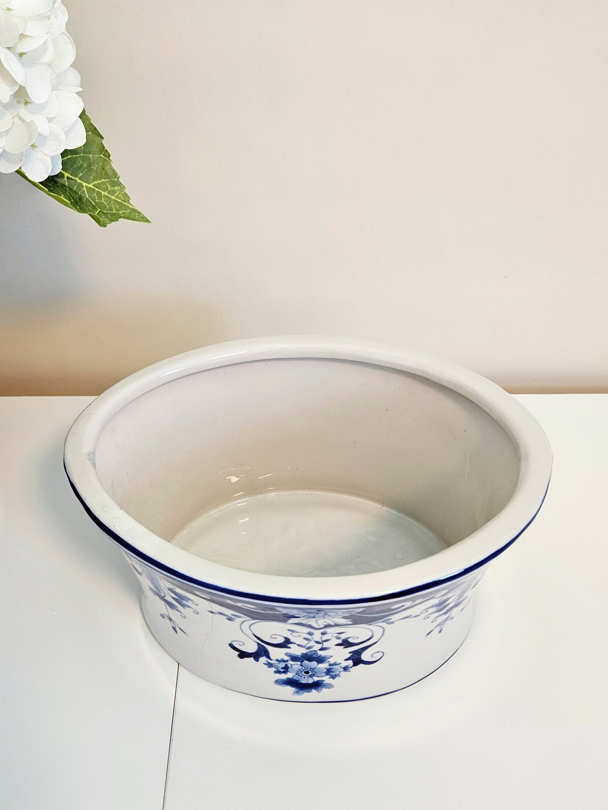 A chinoiserie blue and white planter pot with blue floral patterns and a blue rim, shown on a white surface against a beige wall with a decorative flower in the top-left corner.