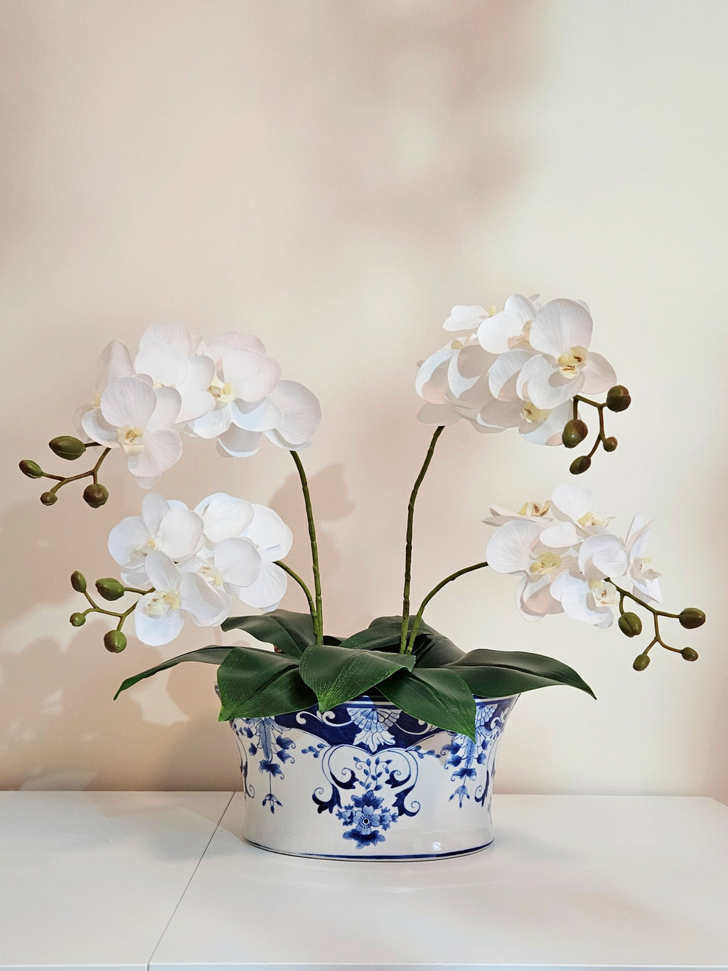 White artificial Phalaenopsis orchid flowers with green stems and leaves, arranged in a blue and white patterned ceramic pot on a white surface against a soft beige background.