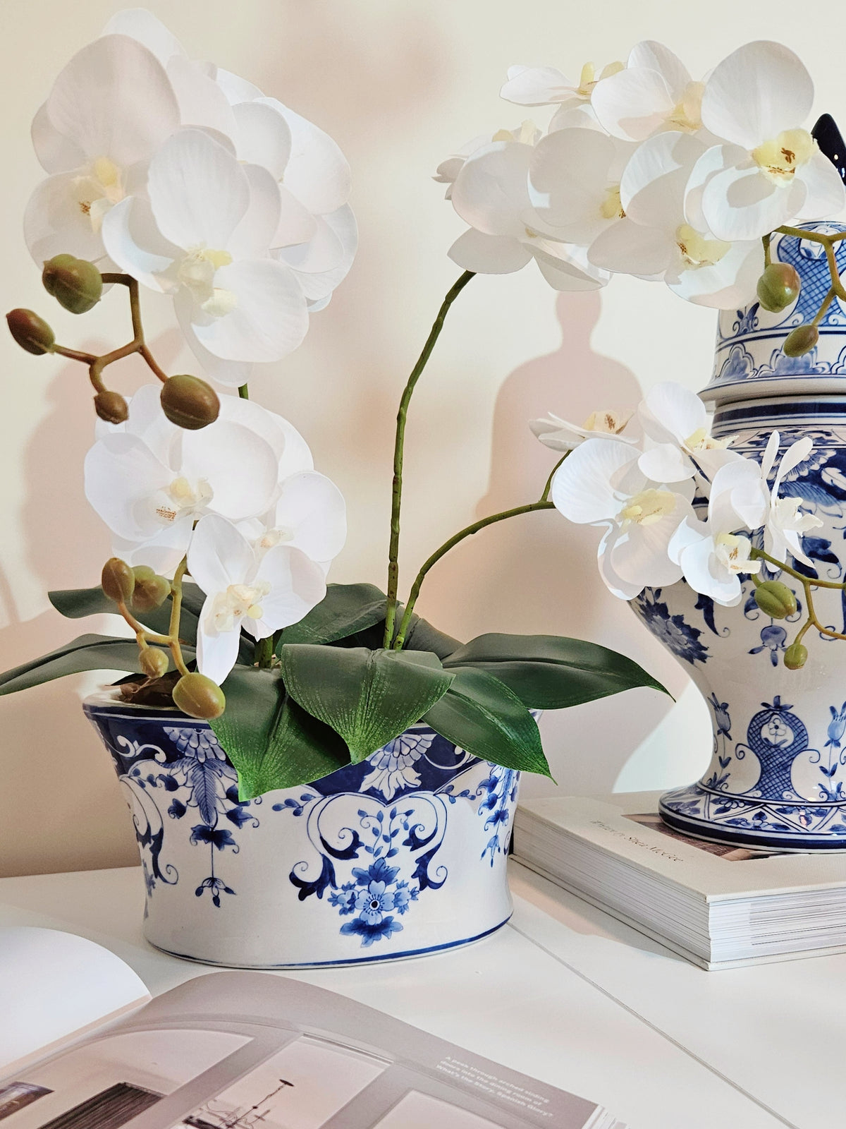 White artificial Phalaenopsis orchid flowers with green leaves in a blue and white porcelain planter pot, styled on a white tabletop next to stacked blue and white ceramic jars and books.