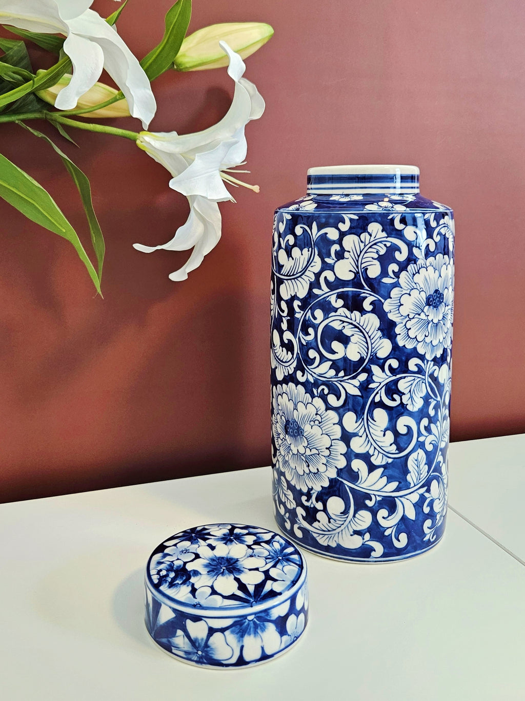 A blue and white floral porcelain jar with its matching lid placed in front, set on a white surface against a terracotta-colored background with white lilies in the upper left.