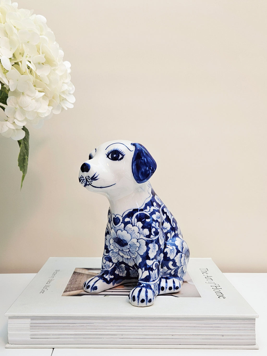 A ceramic dog figurine with a blue and white floral pattern in chinoiserie style sitting on top of a white book with a vase of white flowers in the background.