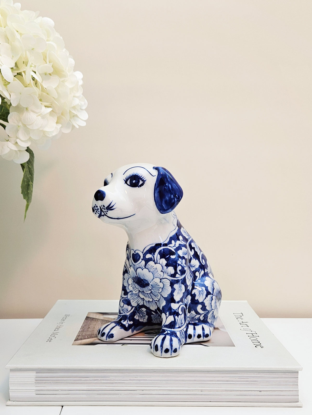 A ceramic dog figurine with a blue and white floral pattern in chinoiserie style sitting on top of a white book with a vase of white flowers in the background.