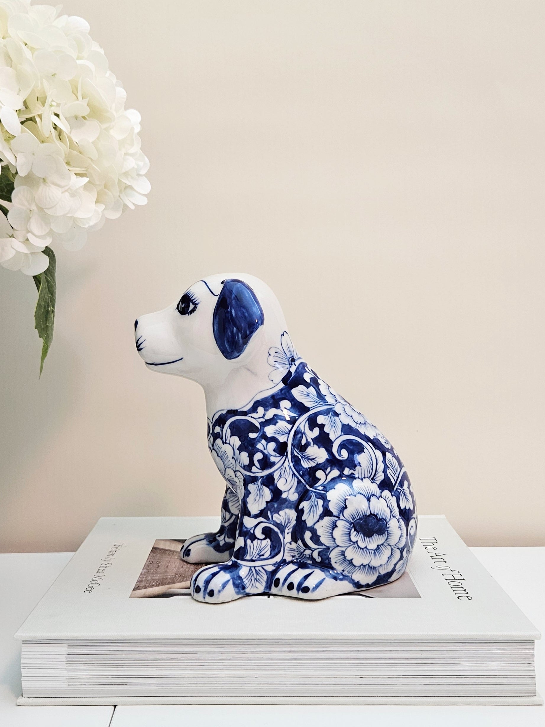 A porcelain dog figurine decorated with a blue and white floral pattern, sitting on top of a white book with white flowers in the background.