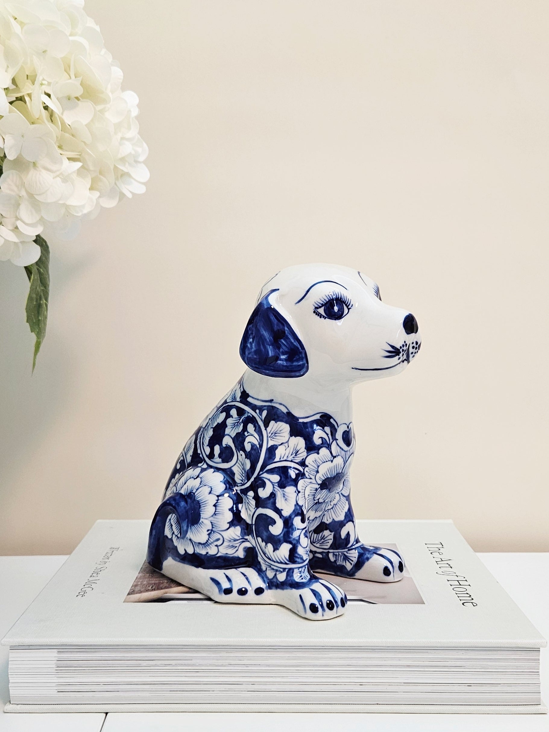 A chinoiserie ceramic dog figurine with a blue and white floral pattern, sitting on top of a white book with a beige background and white flowers in the corner.