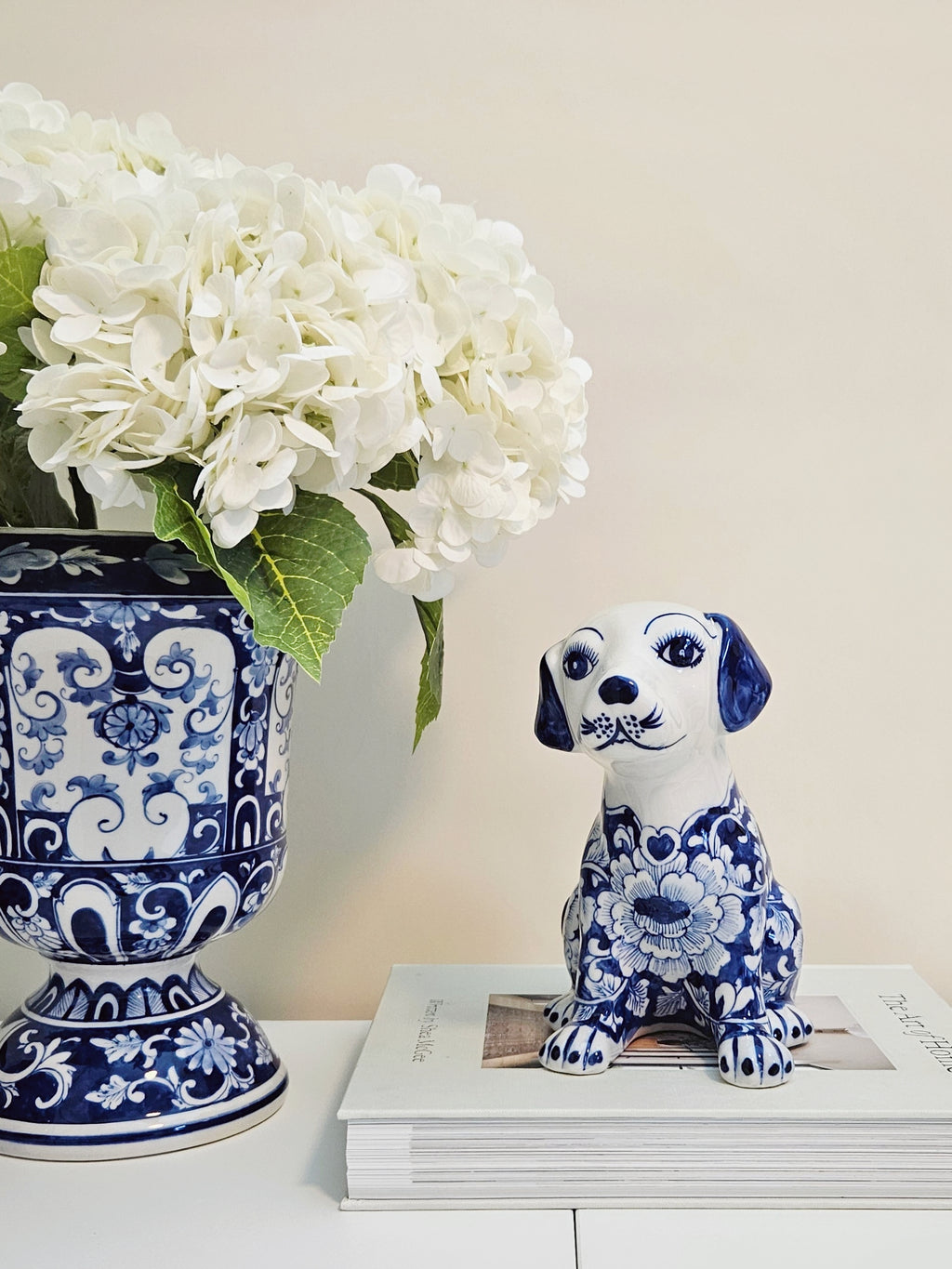 A blue and white porcelain dog figurine with a floral pattern sits on a stack of books next to a matching blue and white floral vase filled with white hydrangeas, set against a warm beige background.