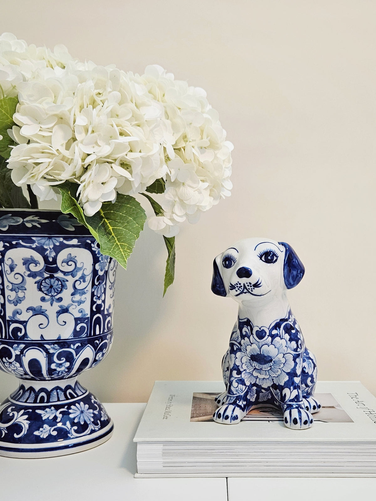 A blue and white porcelain dog figurine with a floral pattern sits on a stack of books next to a matching blue and white floral vase filled with white hydrangeas, set against a warm beige background.