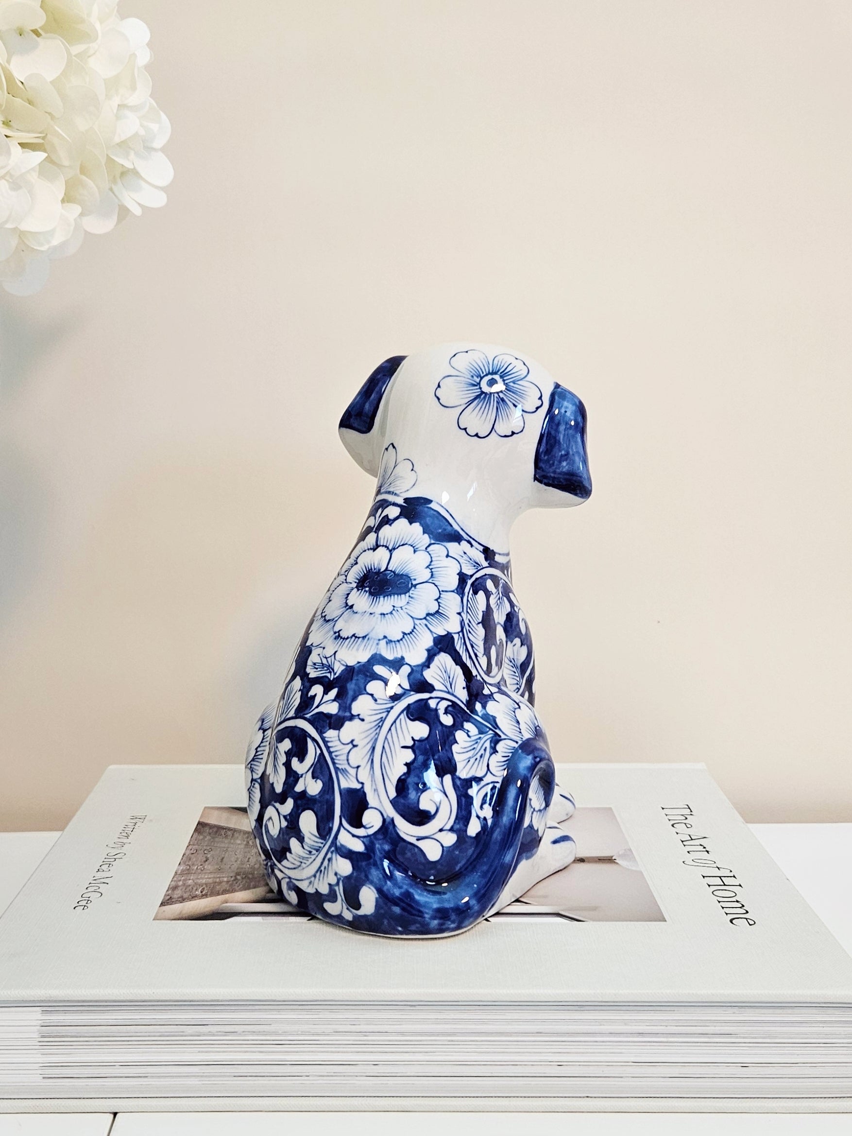 A ceramic dog figurine with a blue and white floral pattern, chinoiserie style, shown from the back sitting on a stack of books against a light beige background.
