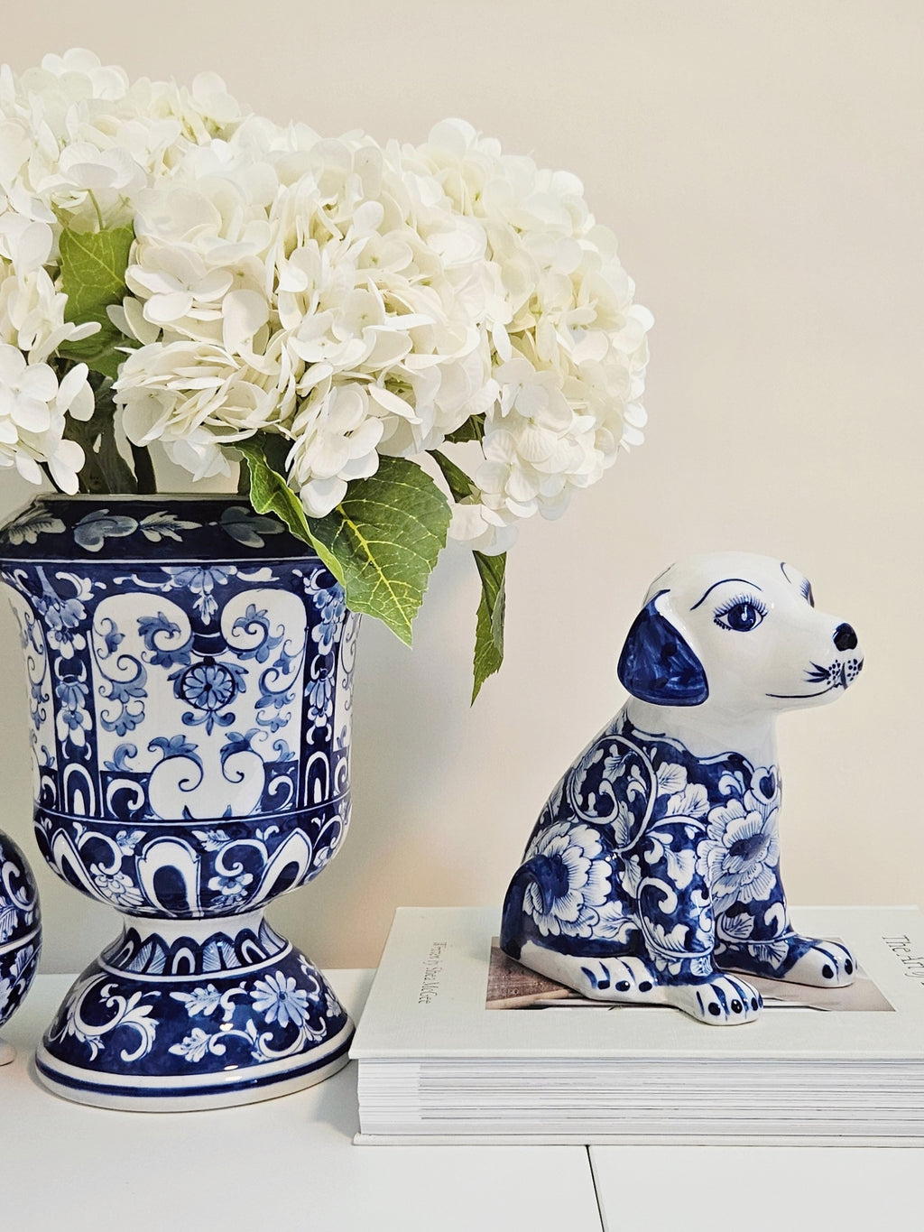 A blue and white porcelain dog figurine with floral patterns sits on a stack of white books next to a matching blue and white floral vase filled with white flowers, set against a warm beige background.