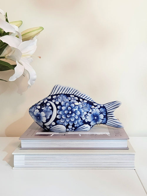 A blue and white porcelain floral fish decorative figurine resting on top of two stacked books against a warm off-white background, with white lily flowers visible in the upper left corner.