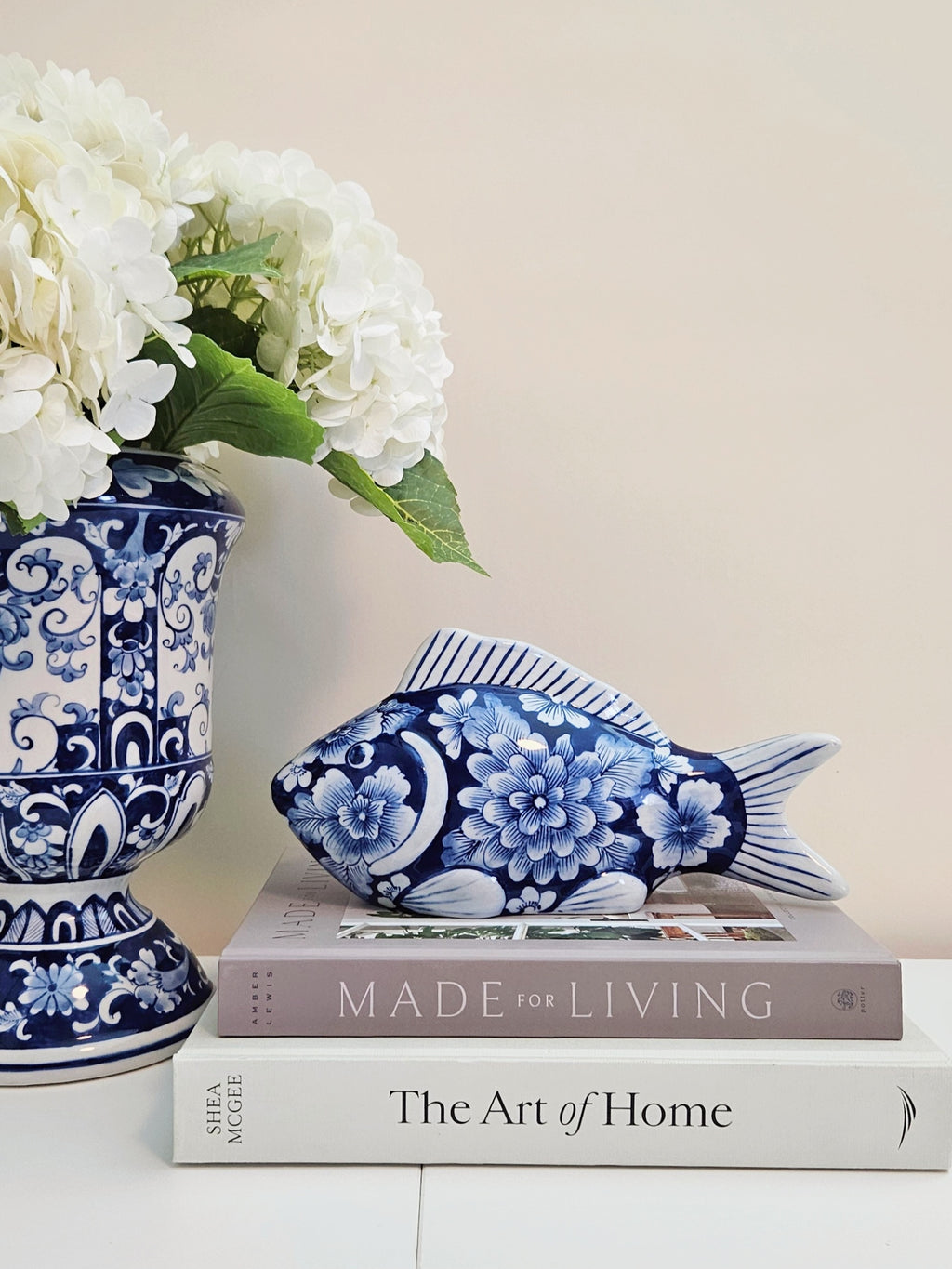 A blue and white chinoiserie floral fish-shaped decorative figurine resting on stacked books, with a matching blue and white patterned vase of white flowers beside it against a plain beige wall.