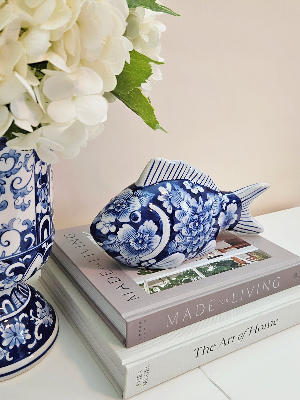 A blue and white porcelain fish figurine with floral patterns, displayed on top of two books next to a blue and white patterned vase with white flowers.