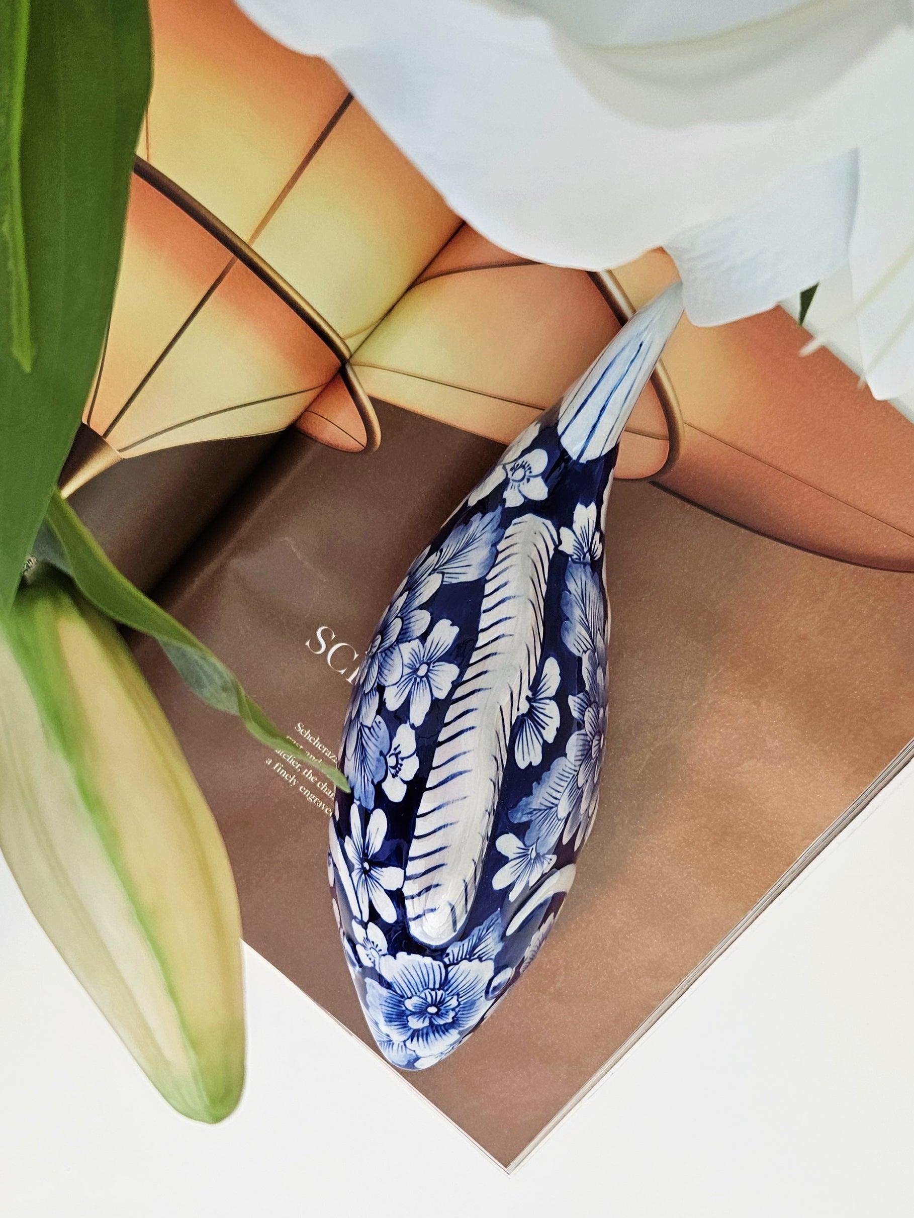 A ceramic decorative fish figurine with a blue and white floral and fish-inspired pattern, styled on a brown book with green leaves and white flowers in the background.