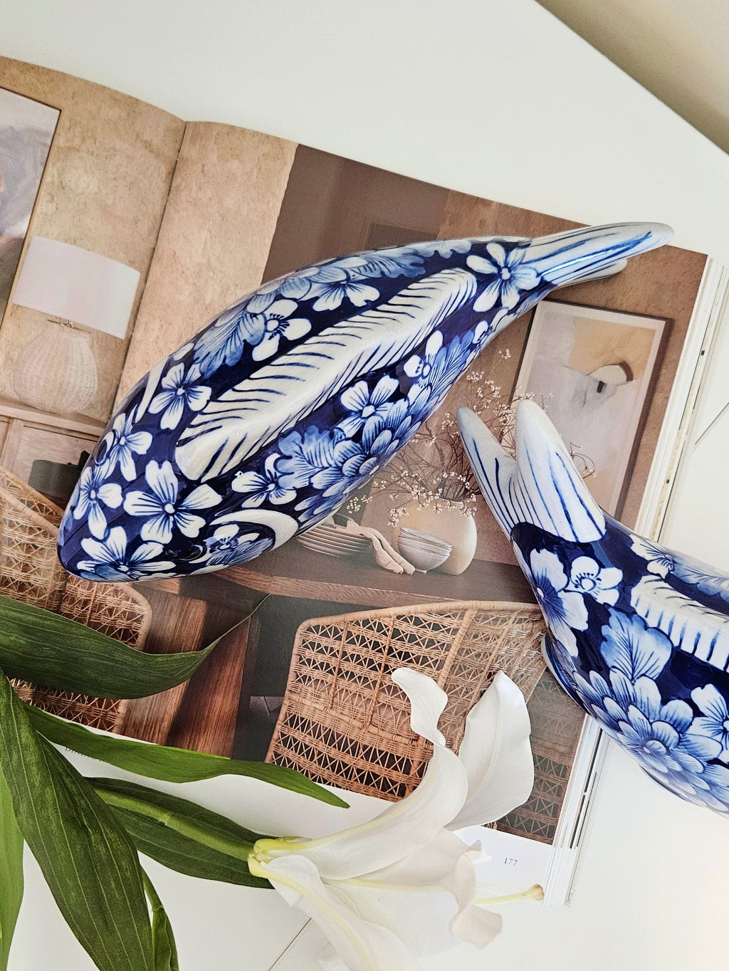 Two ceramic decorative fish sculptures with blue and white floral patterns, displayed on a styled tabletop with an open book and a white lily flower.
