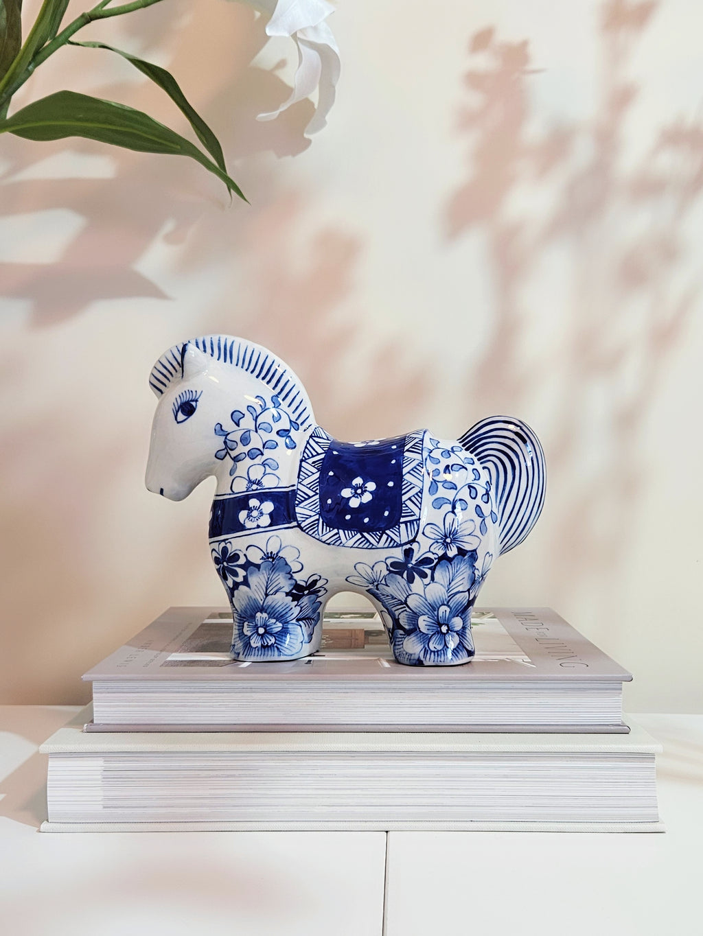 A blue and white hand-painted ceramic horse figurine decorated with floral patterns, displayed on top of two stacked books against a warm beige wall with soft leaf shadows.