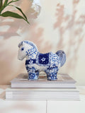 A blue and white hand-painted ceramic horse figurine decorated with floral patterns, displayed on top of two stacked books against a warm beige wall with soft leaf shadows.