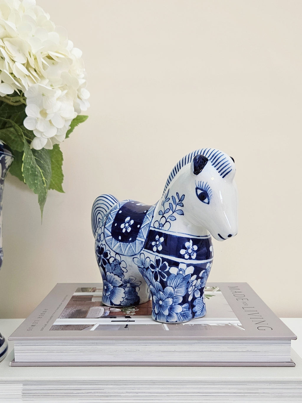 A blue and white ceramic horse figurine with floral patterns, displayed on top of a book with a vase of white flowers in the background.