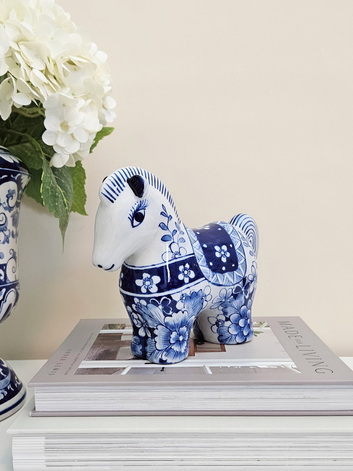 A blue and white ceramic horse figurine with floral patterns, displayed on top of a magazine next to a matching blue and white vase with white flowers.