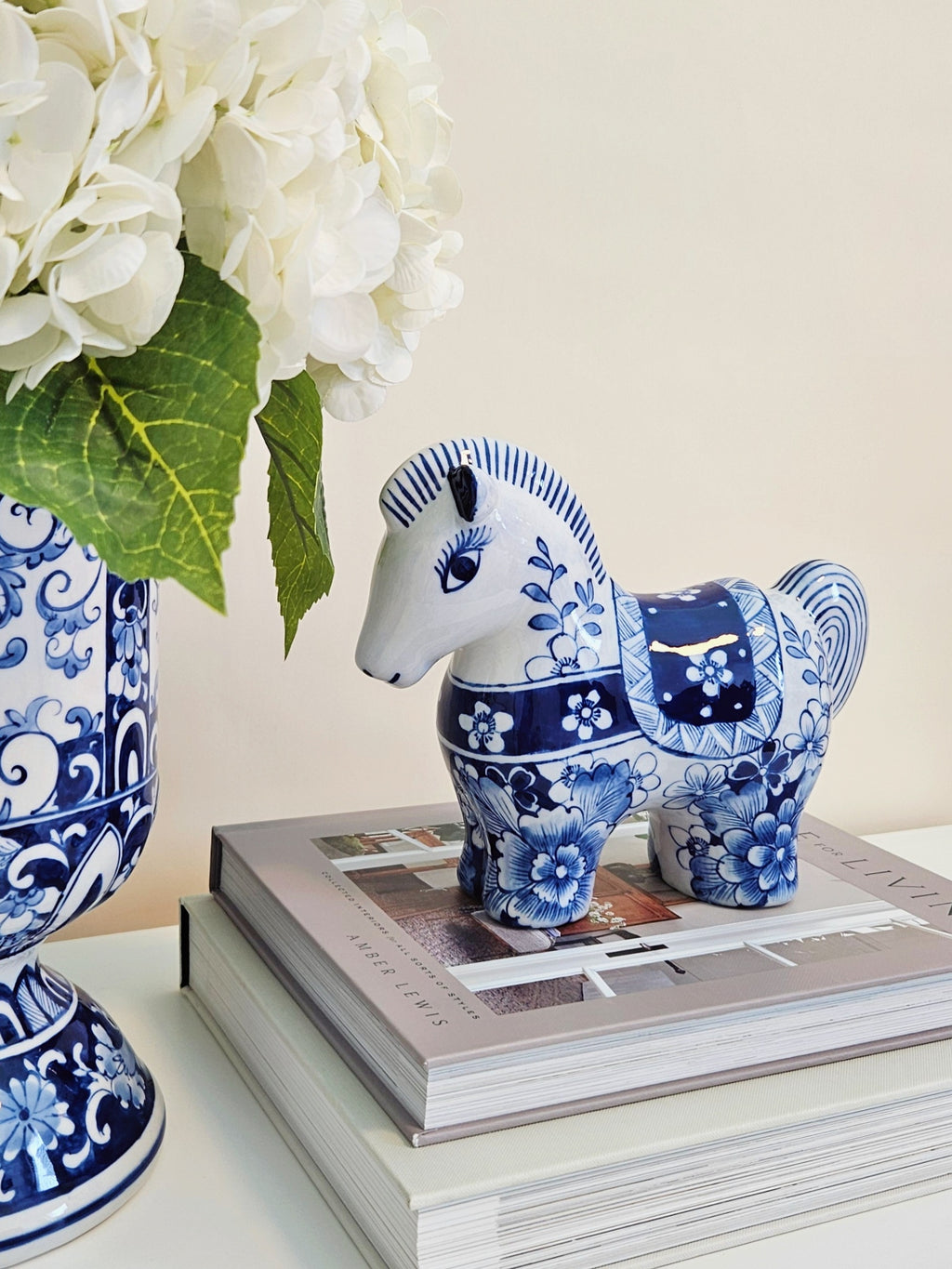 A blue and white patterned ceramic horse figurine displayed on top of stacked books next to a matching blue and white floral vase with white flowers.