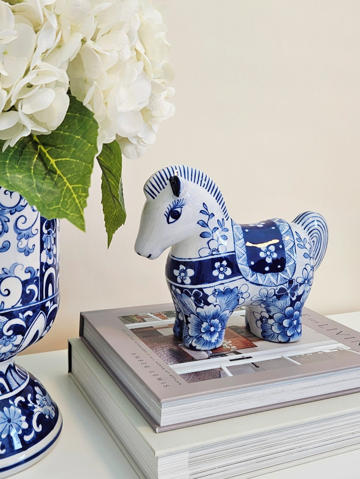 A blue and white patterned ceramic horse figurine displayed on top of stacked books next to a matching blue and white floral vase with white flowers.