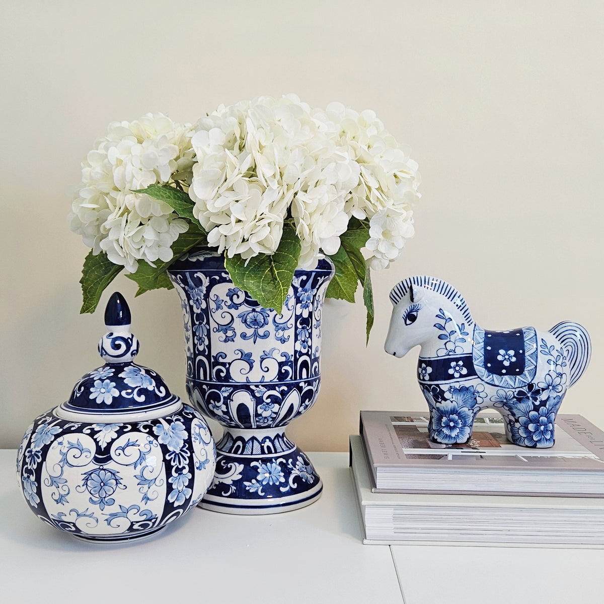 Blue and white porcelain decorative pieces on a white surface against a beige wall, including a lidded jar, a pedestal vase filled with white flowers, and a horse figurine on stacked books.